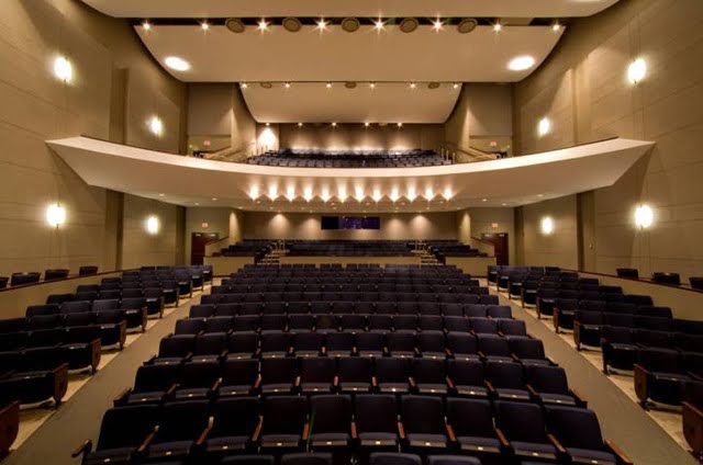 Empty auditorium with rows of dark seats, balcony level, and stage lighting, viewed from the stage facing the audience area.